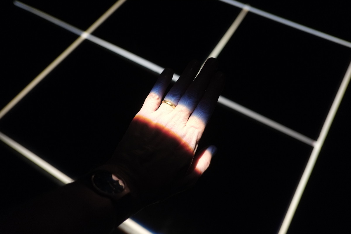 A hand lit by a strip of light cutting across. A white grid on a black floor behind.