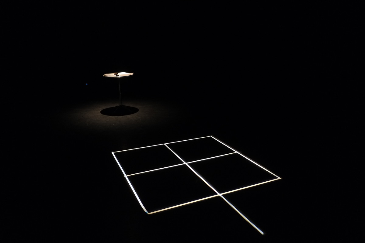 Black space with white grid on floor, and a dimly illuminated round table at rear