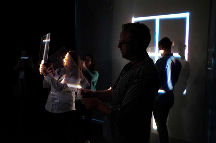 Darkened room with people holding up small perspex screens in narrow cuts of light