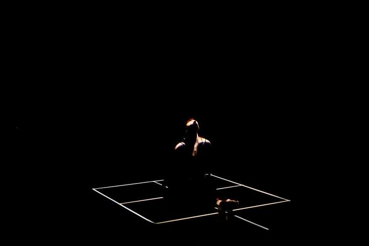 Grid of white lines in a dark space. Parts of the performer's body are glimpsed in narroew cuts of light