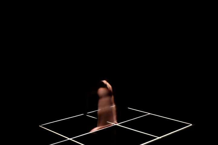 Grid of white lines in a dark space. Parts of the performer's body are glimpsed in narroew cuts of light