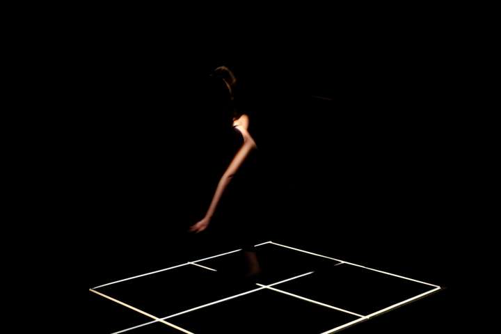 Grid of white lines in a dark space. Parts of the performer's body are glimpsed in narroew cuts of light