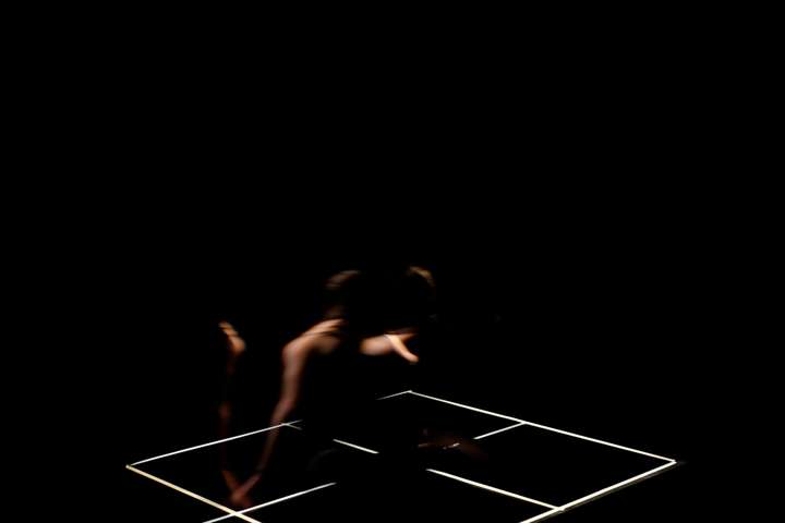 Grid of white lines in a dark space. Parts of the performer's body are glimpsed in narroew cuts of light